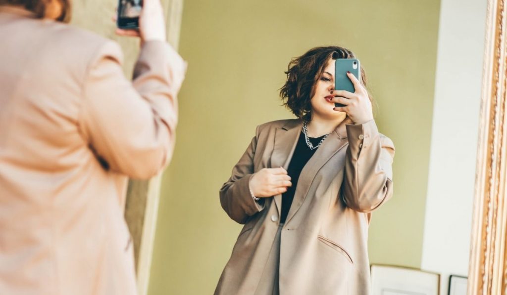 plus size woman taking a picture while trying on a brown coat - ee220321