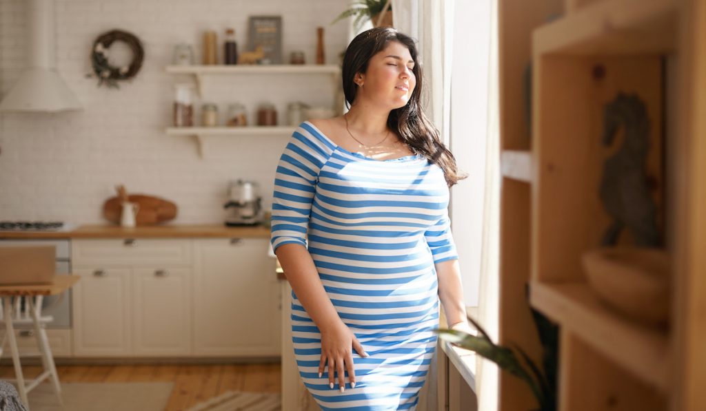 woman wearing blue and white horizontal stripes dress by the window feeling the sunlight