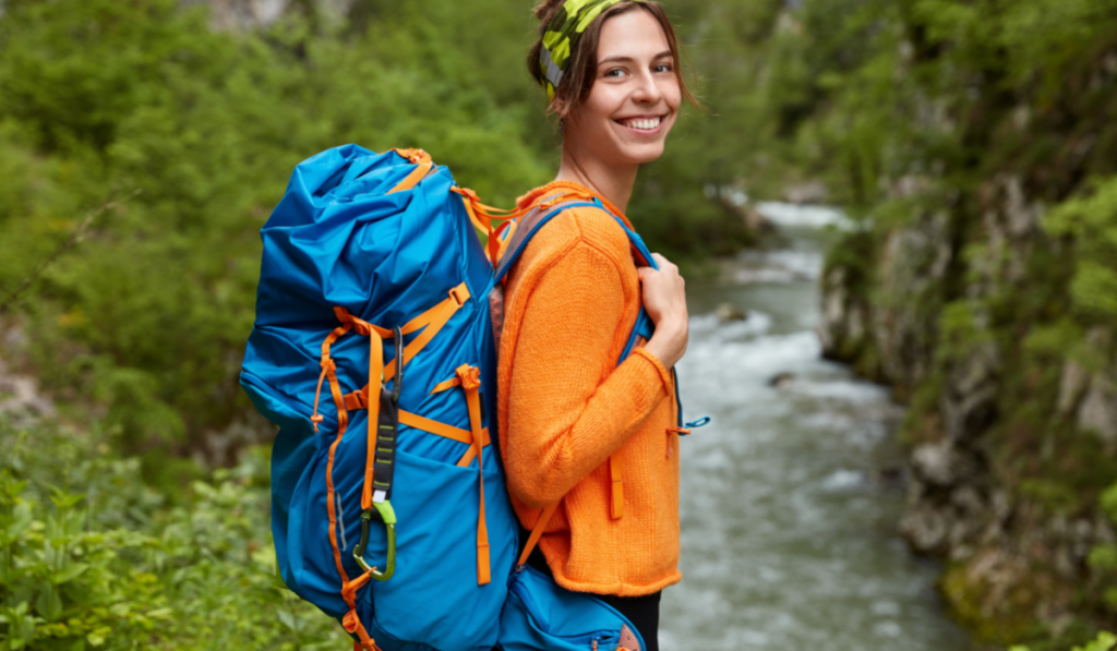 cheerful woman trekking in orange jumper carrying a blue backpack 